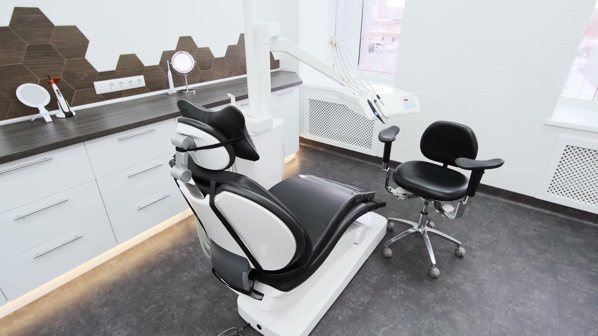 A quiet, empty treatment room: a dark dental chair, white cabinets, a large wall clock and soft light through window blinds