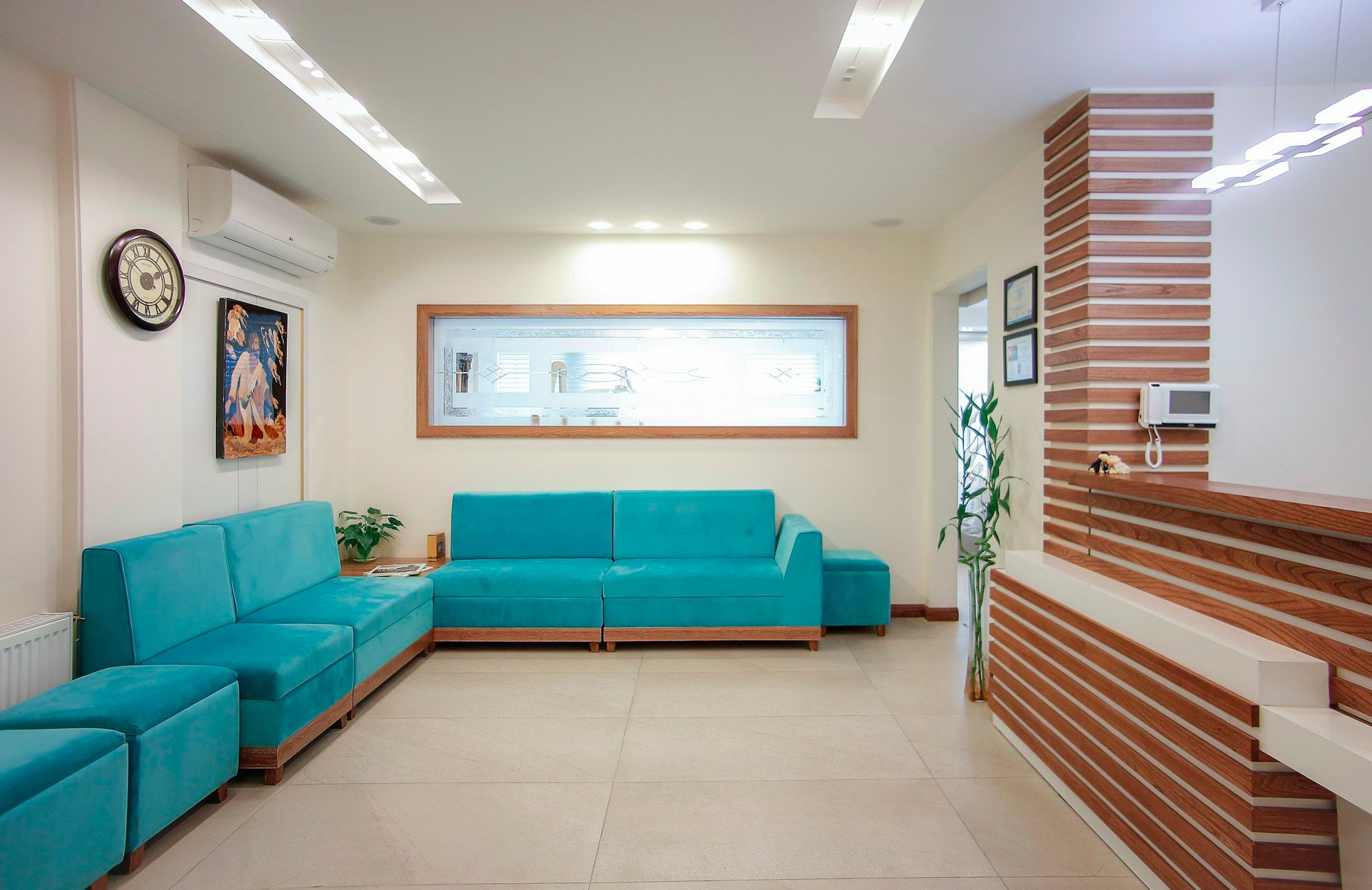 A waiting room with a long teal sofa, a plant and a window into the hallway